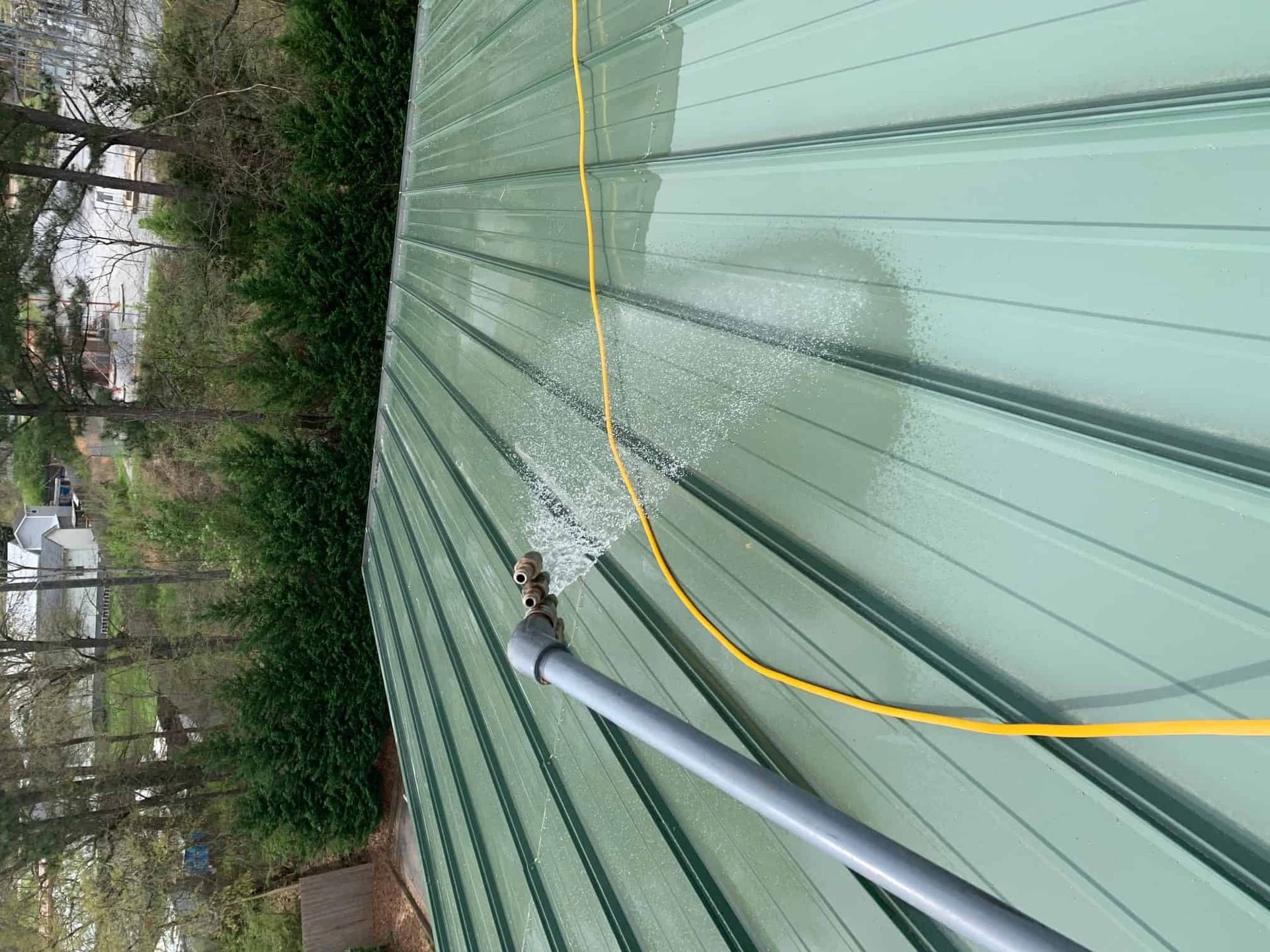 A metal roof is being pressure washed with a spray wand, cleaning a patch of green roofing material.