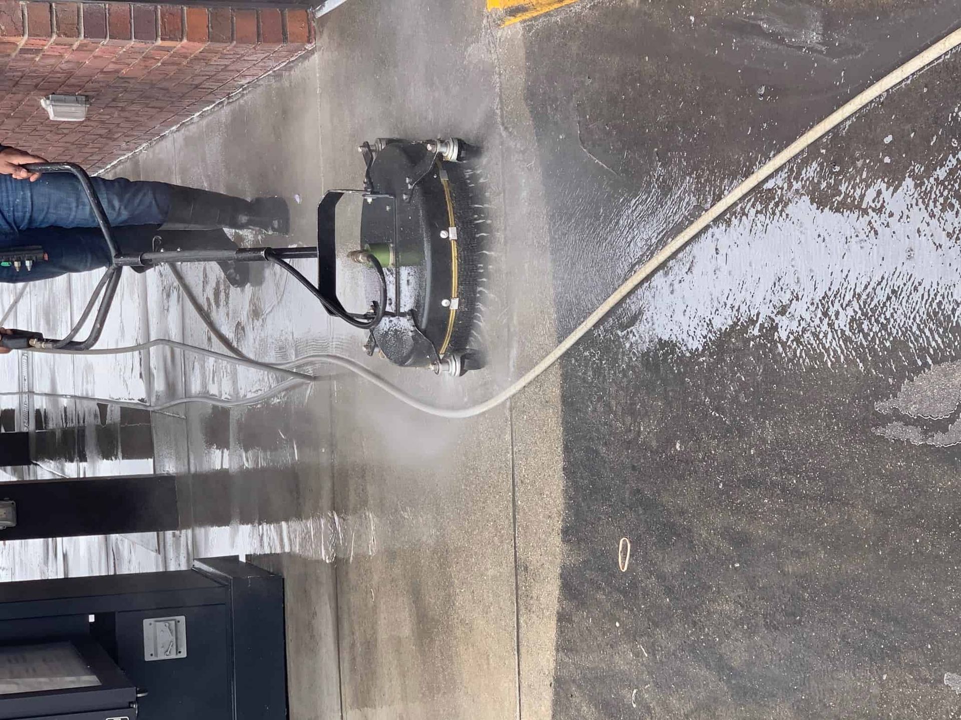 A person uses a circular surface cleaner attached to a pressure washer to clean a concrete sidewalk.