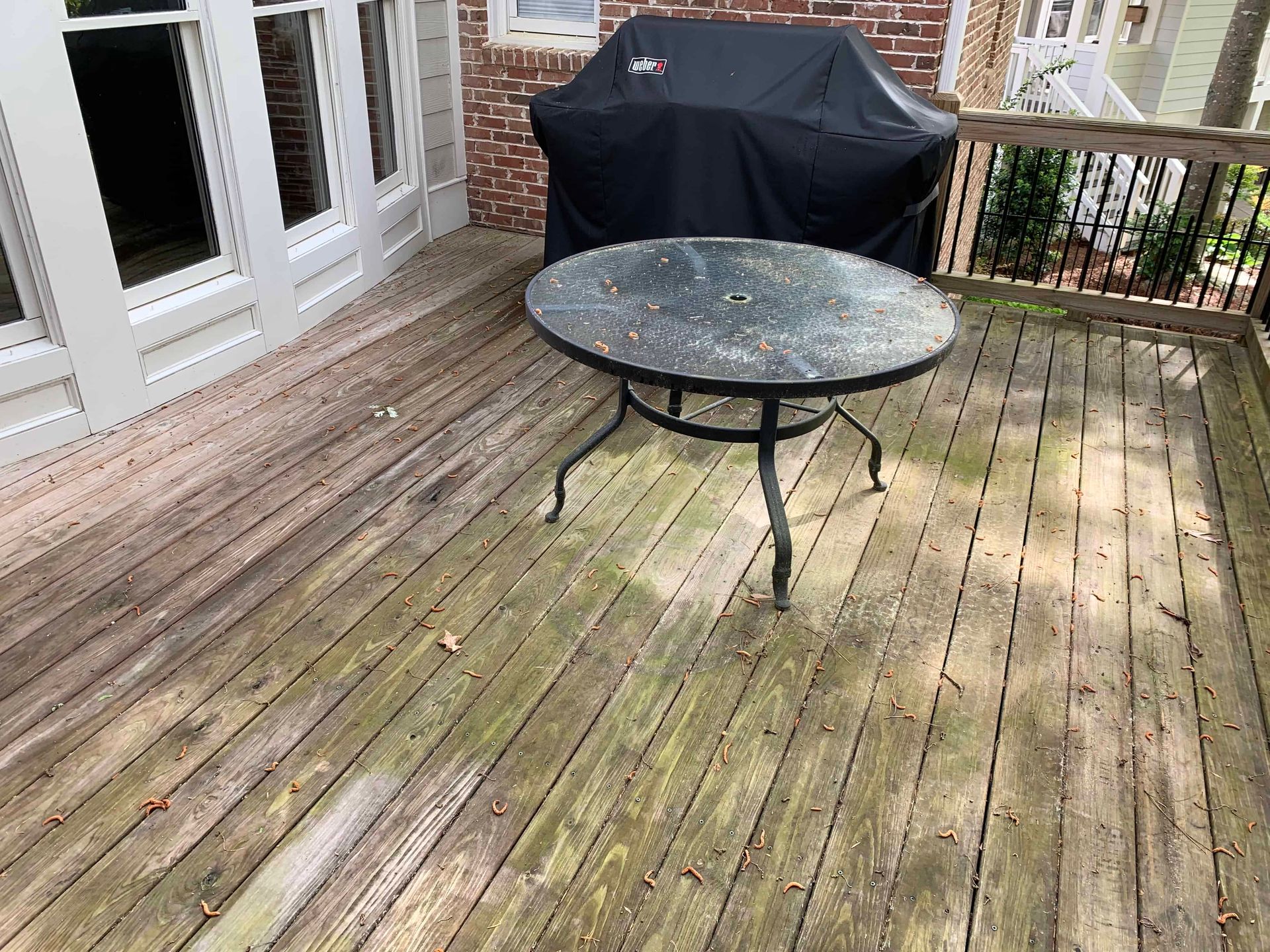 A weathered wooden deck with a round metal table and a covered grill near a brick house wall.