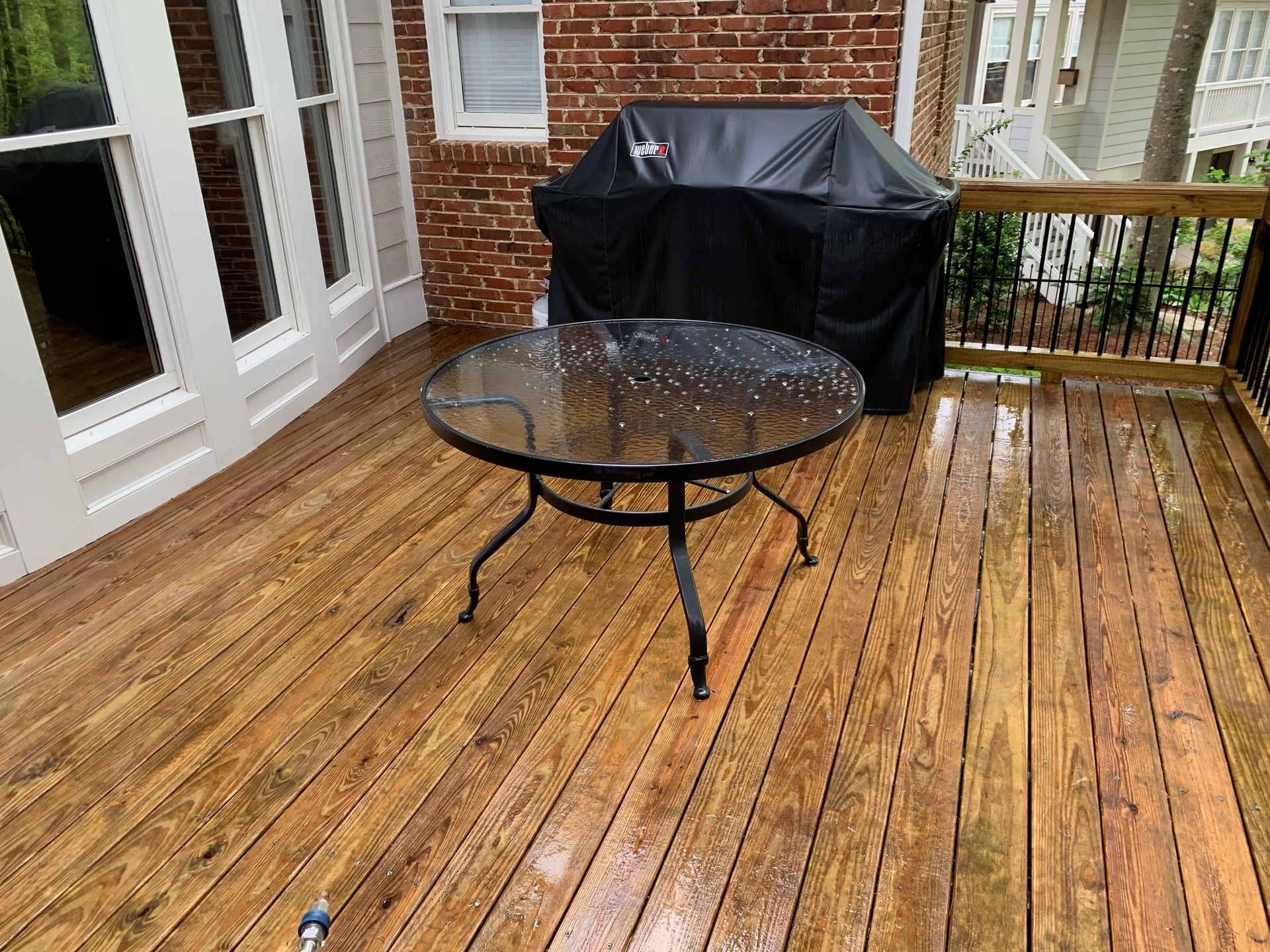 A round glass patio table sits on a wet wooden deck, with a covered grill against a brick wall in the background.