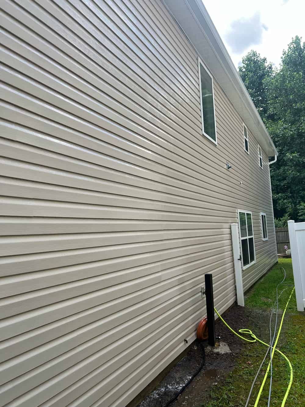 The side view of a two-story beige house with horizontal vinyl siding, two windows, and a black support pole in the yard.