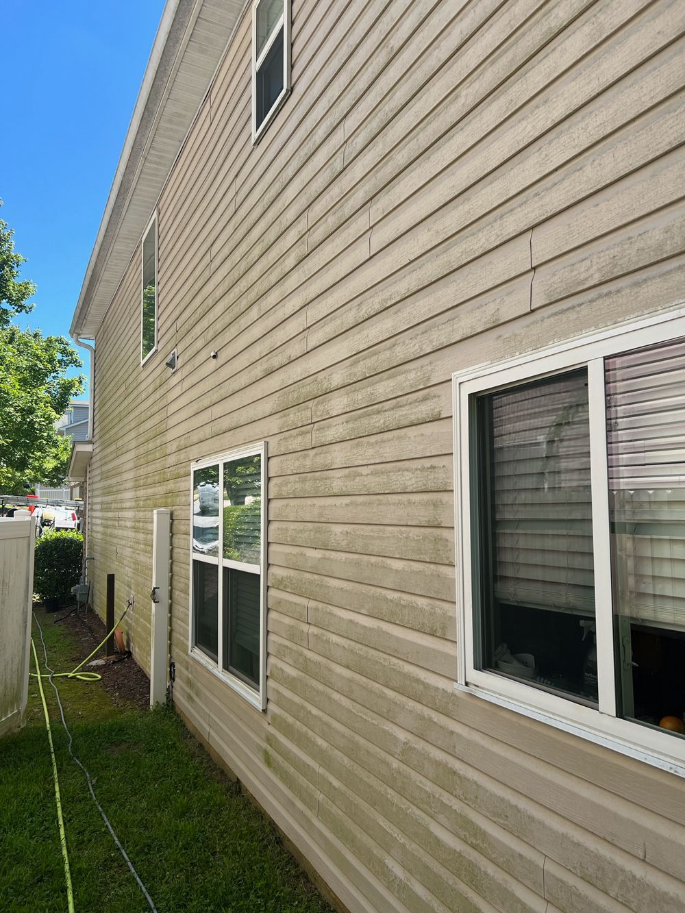 Tan vinyl siding on the side of a house, showing significant dirt and algae buildup, with several windows visible.
