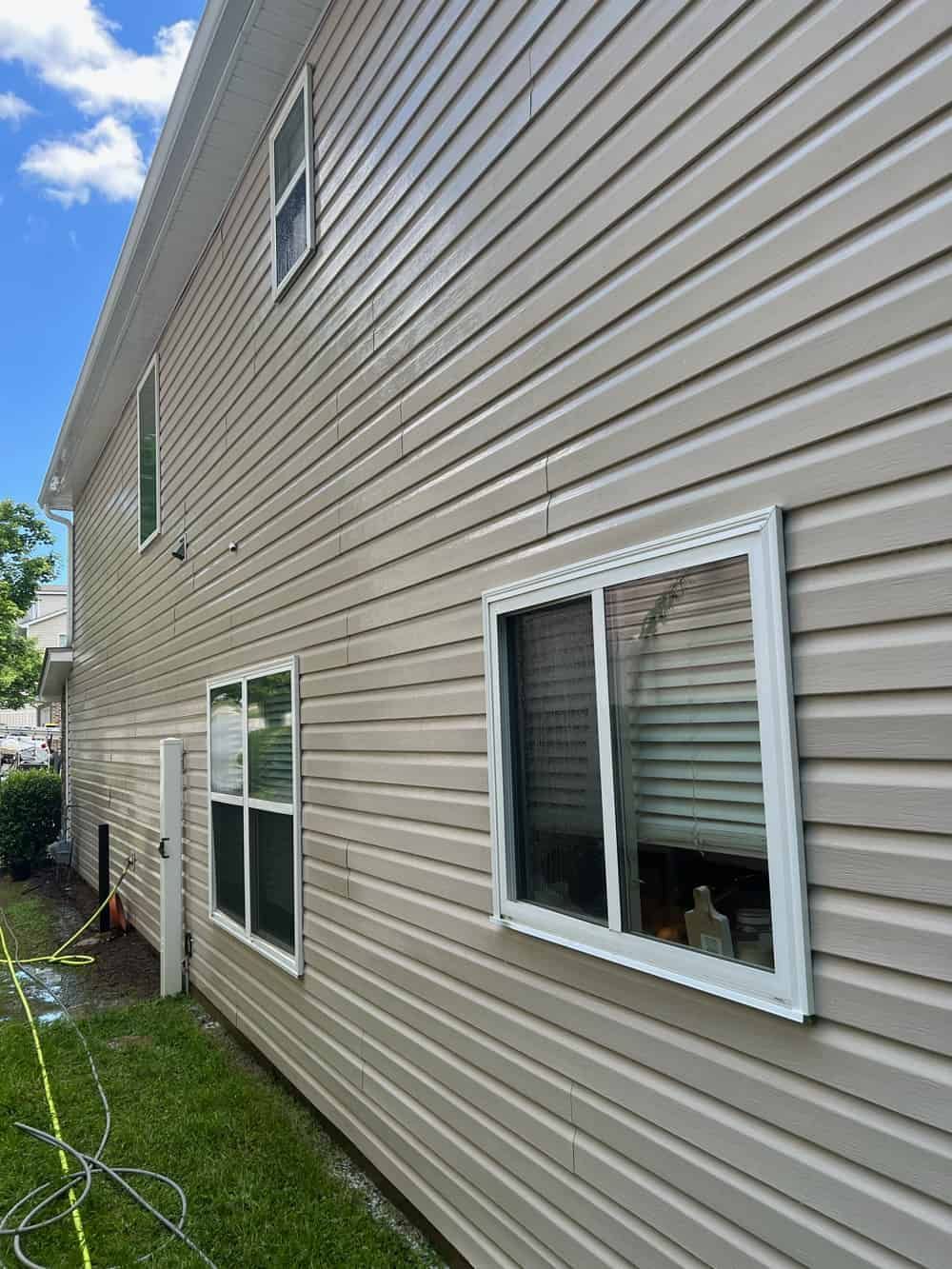 A view of a beige vinyl-sided house exterior with two windows and a green lawn under a sunny, blue sky.