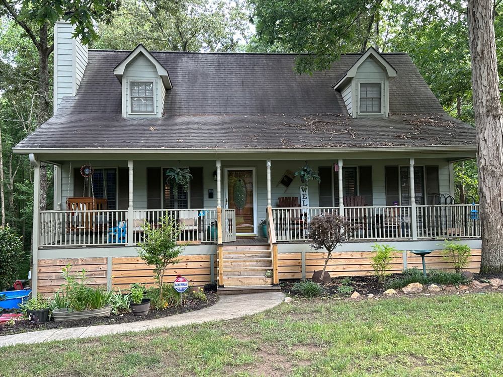 A light green house with a front porch, dormer windows, and dark roof sits in a wooded yard with a stone walkway.