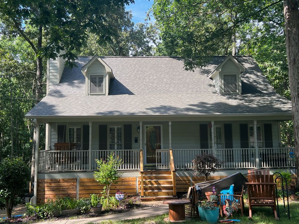 A light blue house with a covered front porch, two dormer windows, and wooden lattice skirting, nestled in a forest.