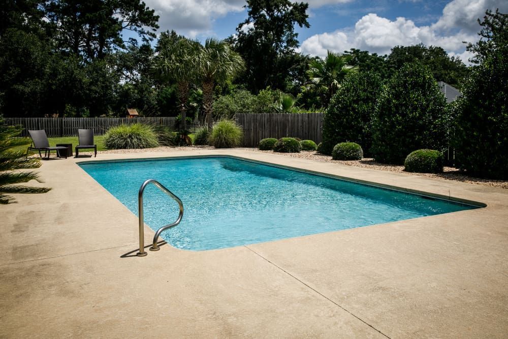 A rectangular backyard swimming pool with blue water and a beige concrete deck, surrounded by trees and shrubs.