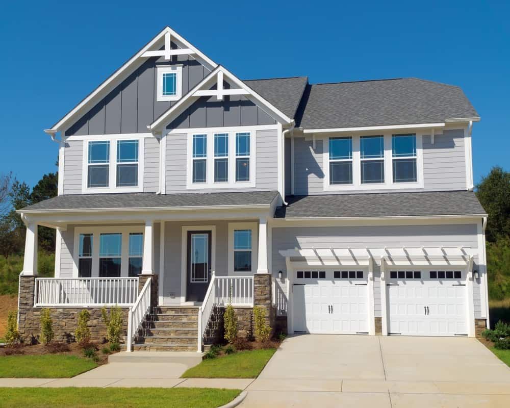 A two-story suburban house with grey siding, a front porch, stone foundation, and a two-car garage under a blue sky.