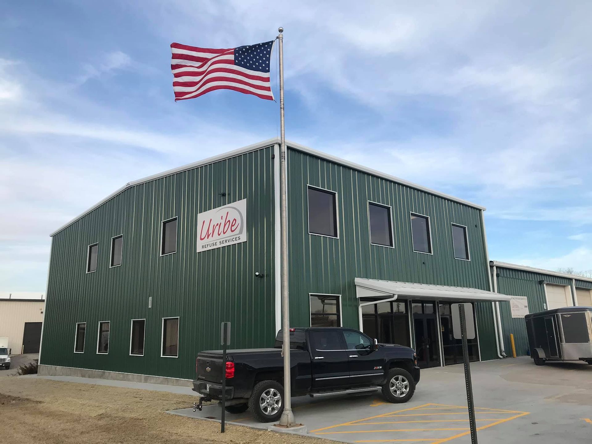 A truck is parked in front of a green building with an american flag flying in the wind.