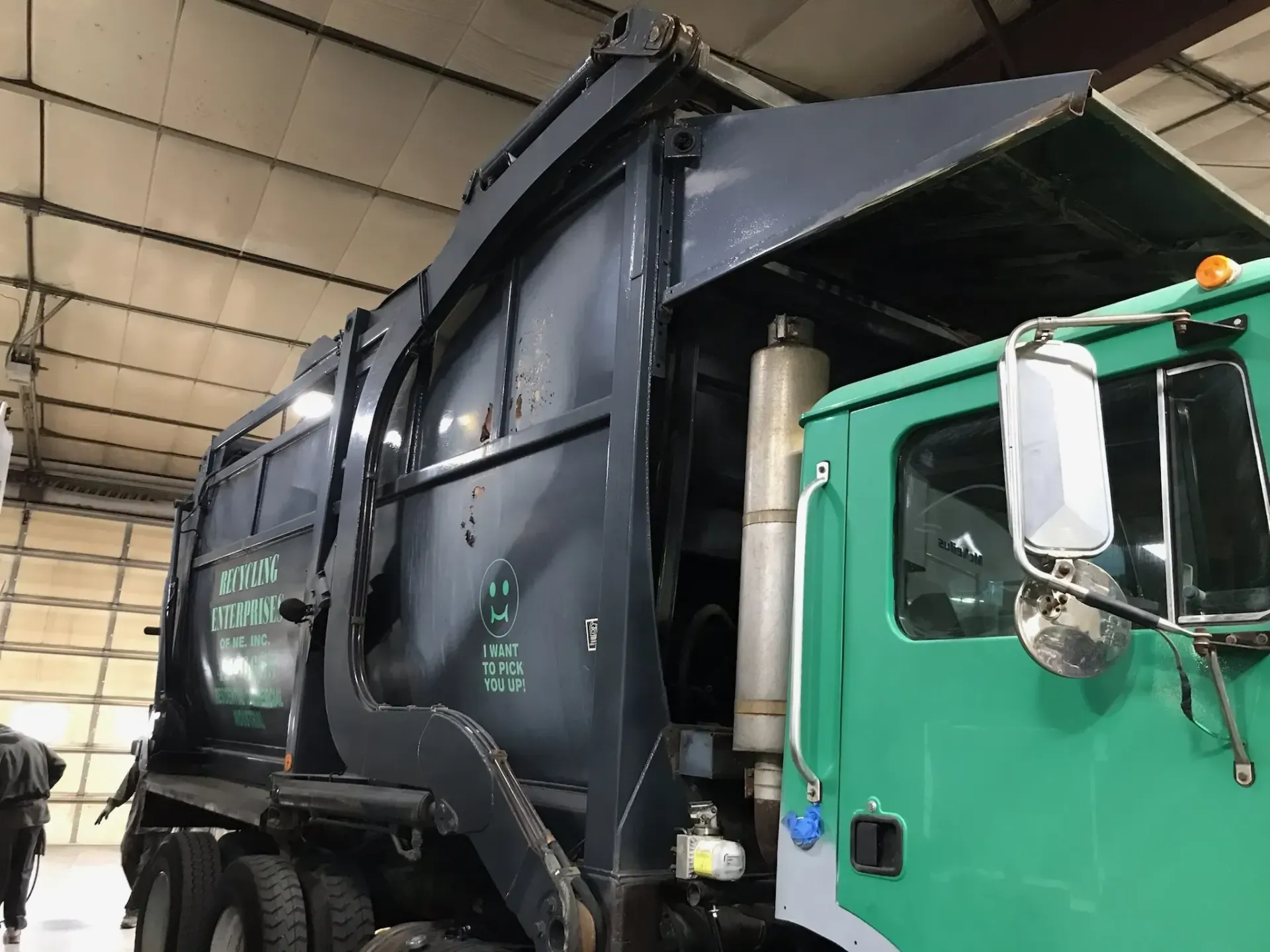 A green garbage truck is parked in a warehouse with its hood open.