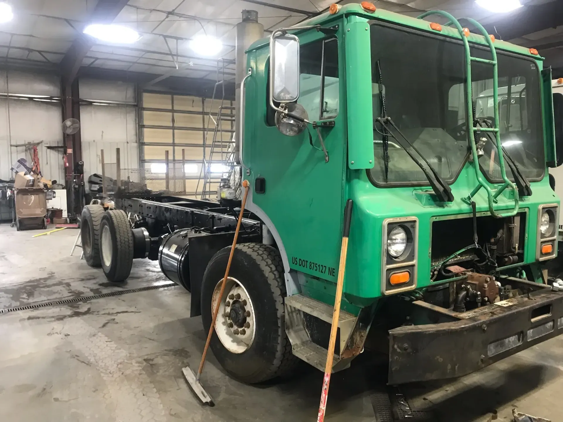 A green garbage truck is sitting in a garage with a broom.