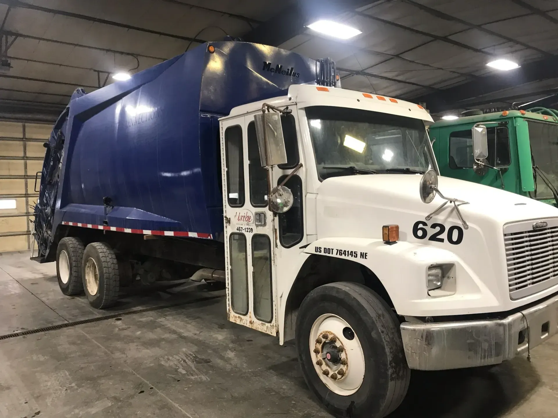 A blue and white garbage truck is parked in a garage.