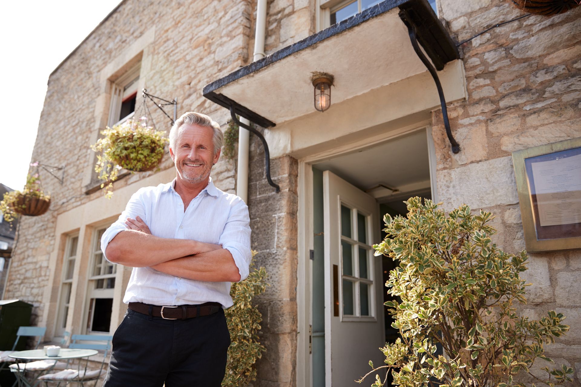 Man with arms crossed stands in front of a stone building with an open door and hanging plants; sunlight.