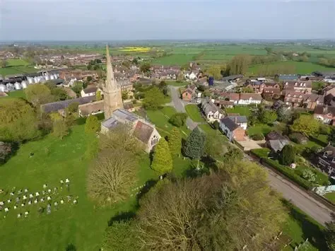 Aerial view of a village church with a tall steeple, surrounded by houses, trees, and green fields.