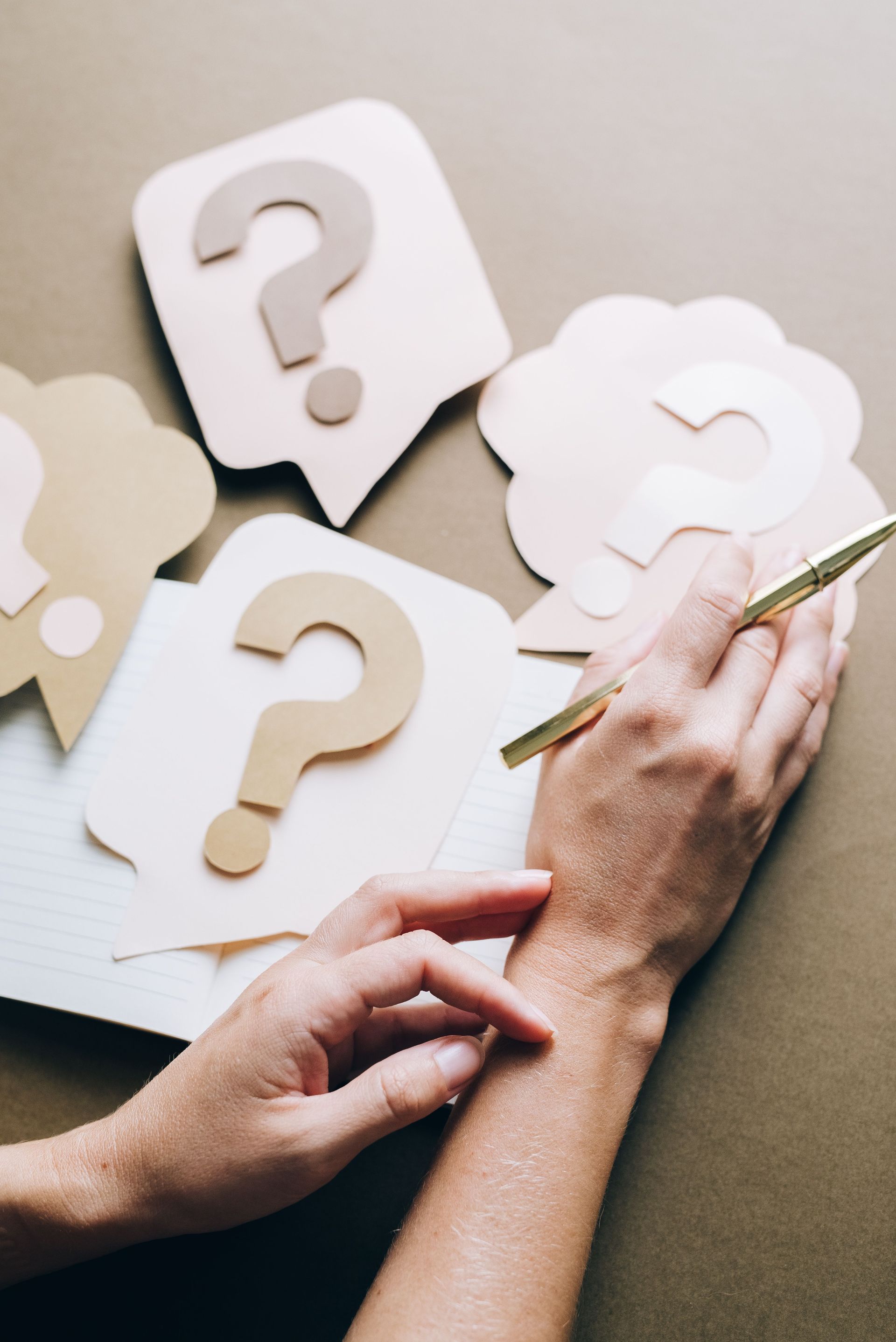 Hands writing on paper next to question-mark speech bubbles with a pen on a beige surface