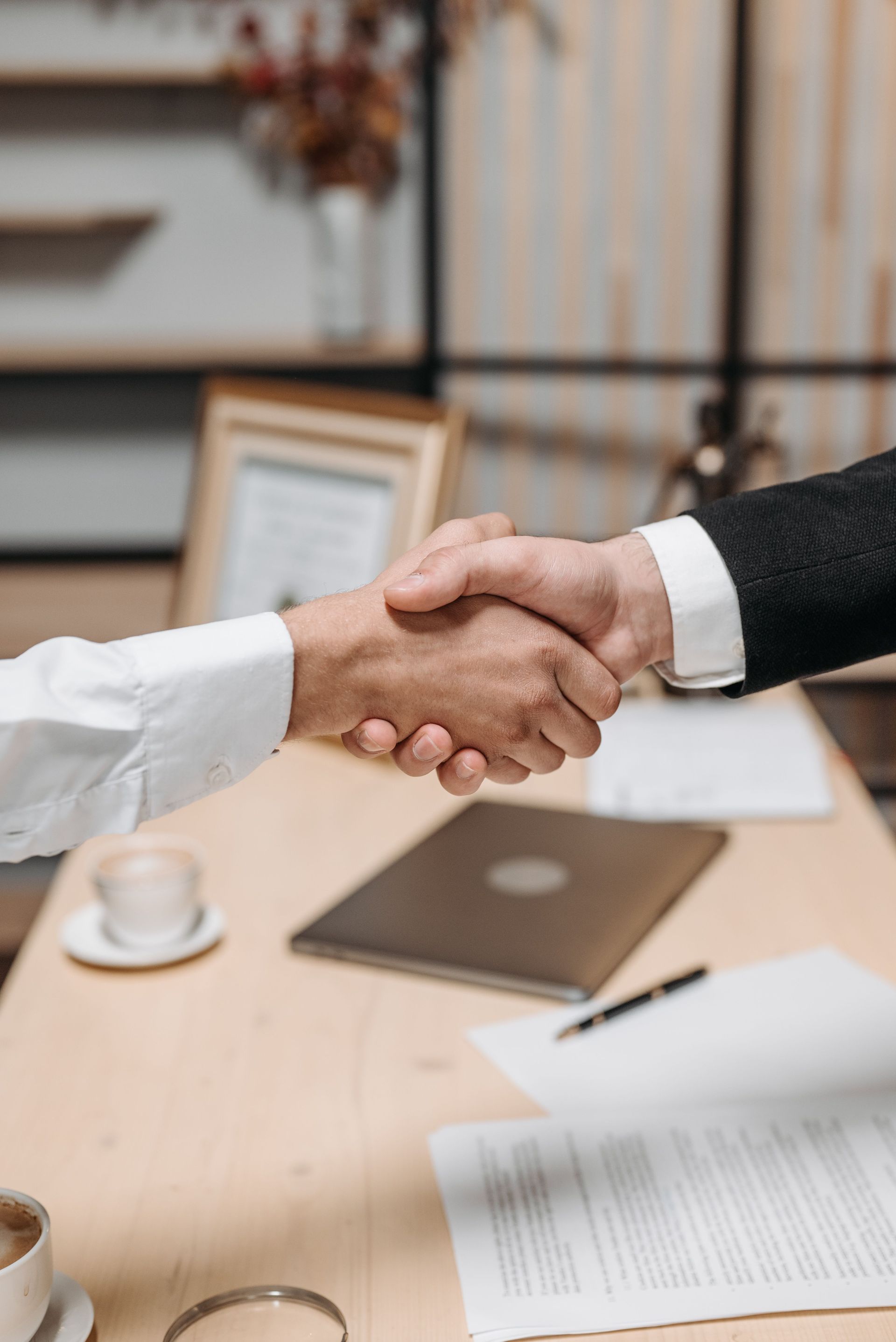 Two people shaking hands across a desk with documents and a laptop in a business meeting