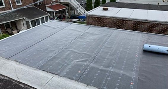 Flat roof covered with grey roofing membrane, roll of material on the right. Houses and foliage in background.