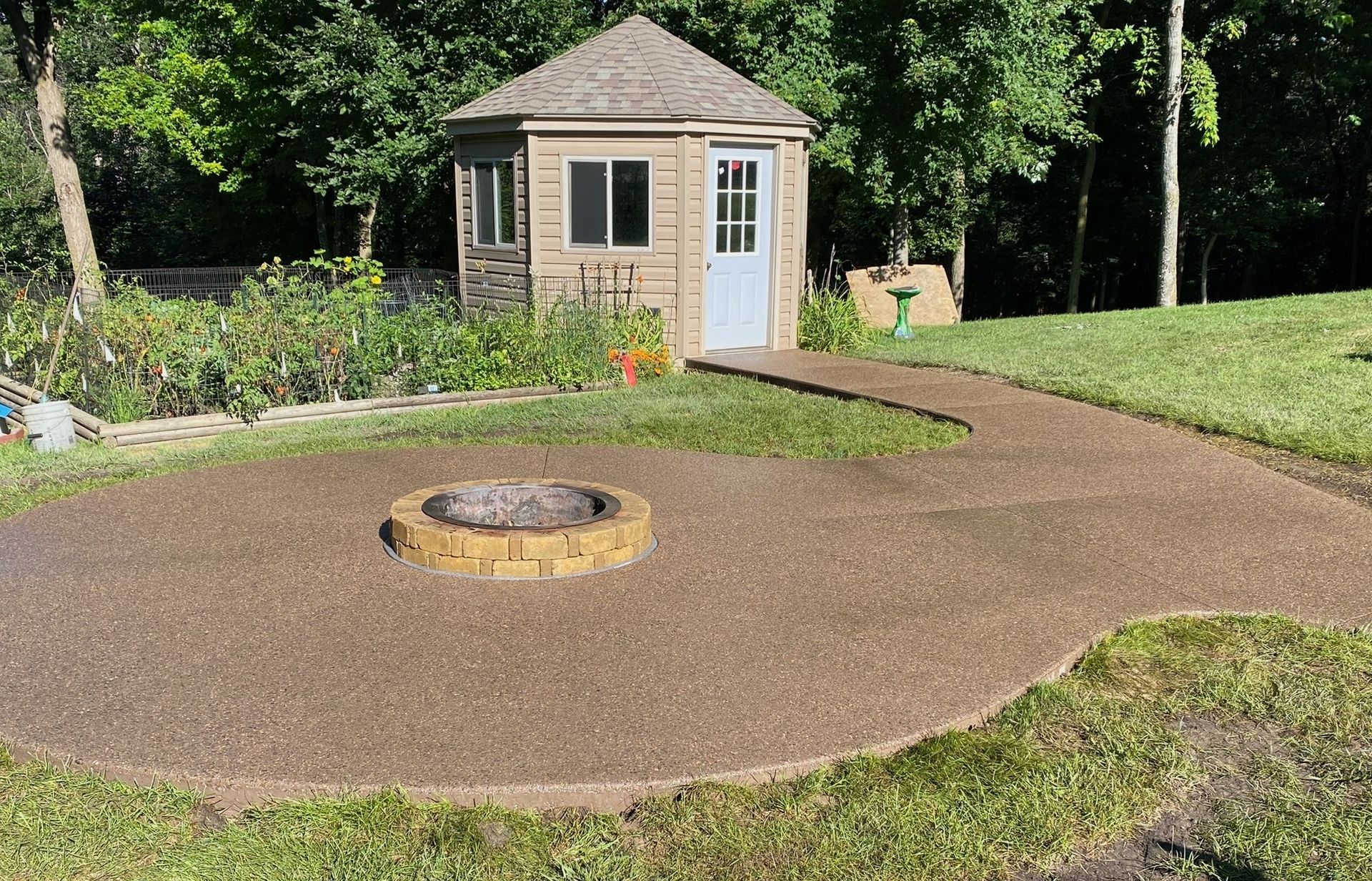 A brown gravel patio with a circular fire pit leads to a small tan shed with a light blue door, set in a sunny backyard.