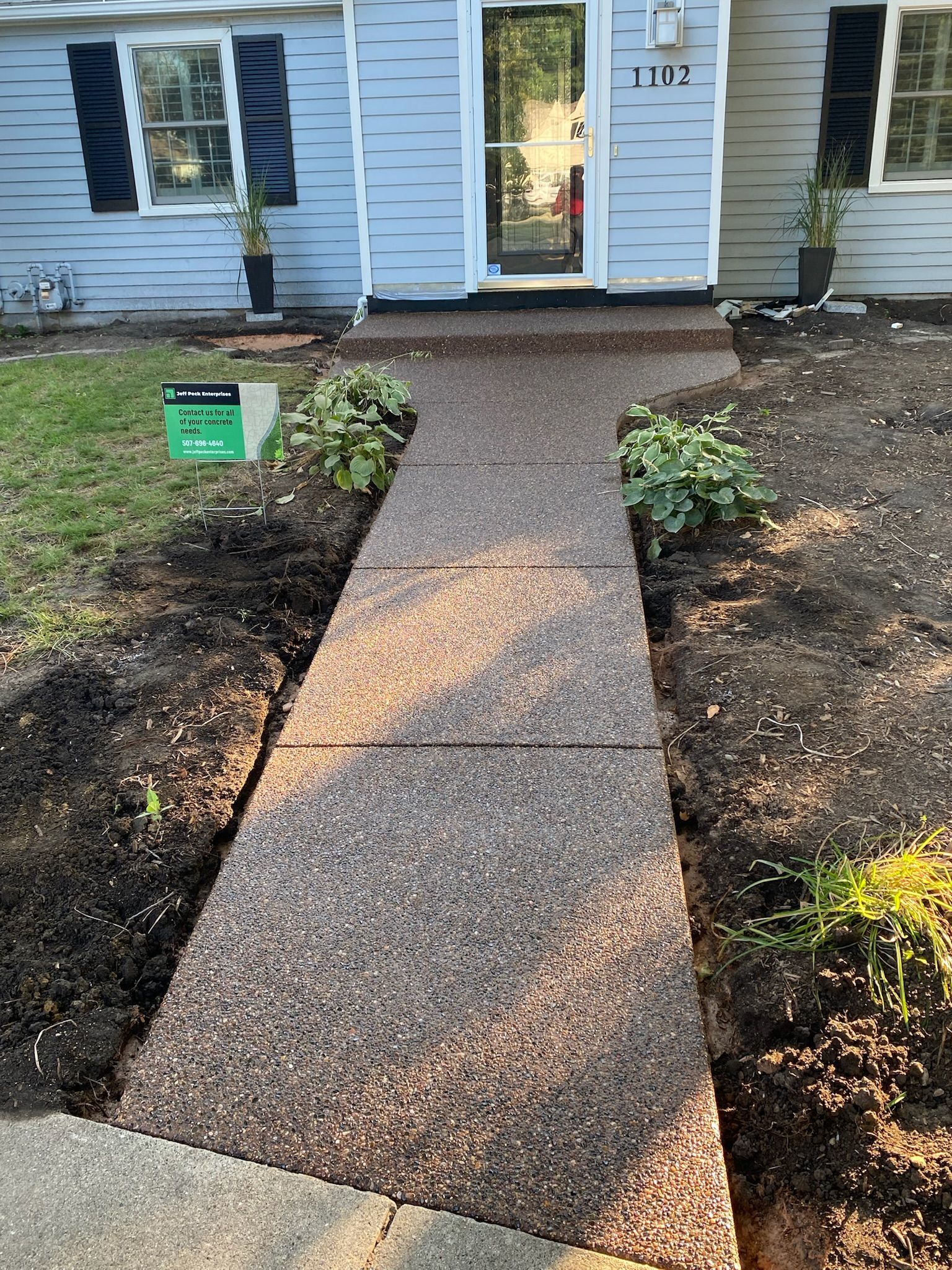 A long, new aggregate concrete walkway leads to the front door of a house with light blue siding and black shutters.
