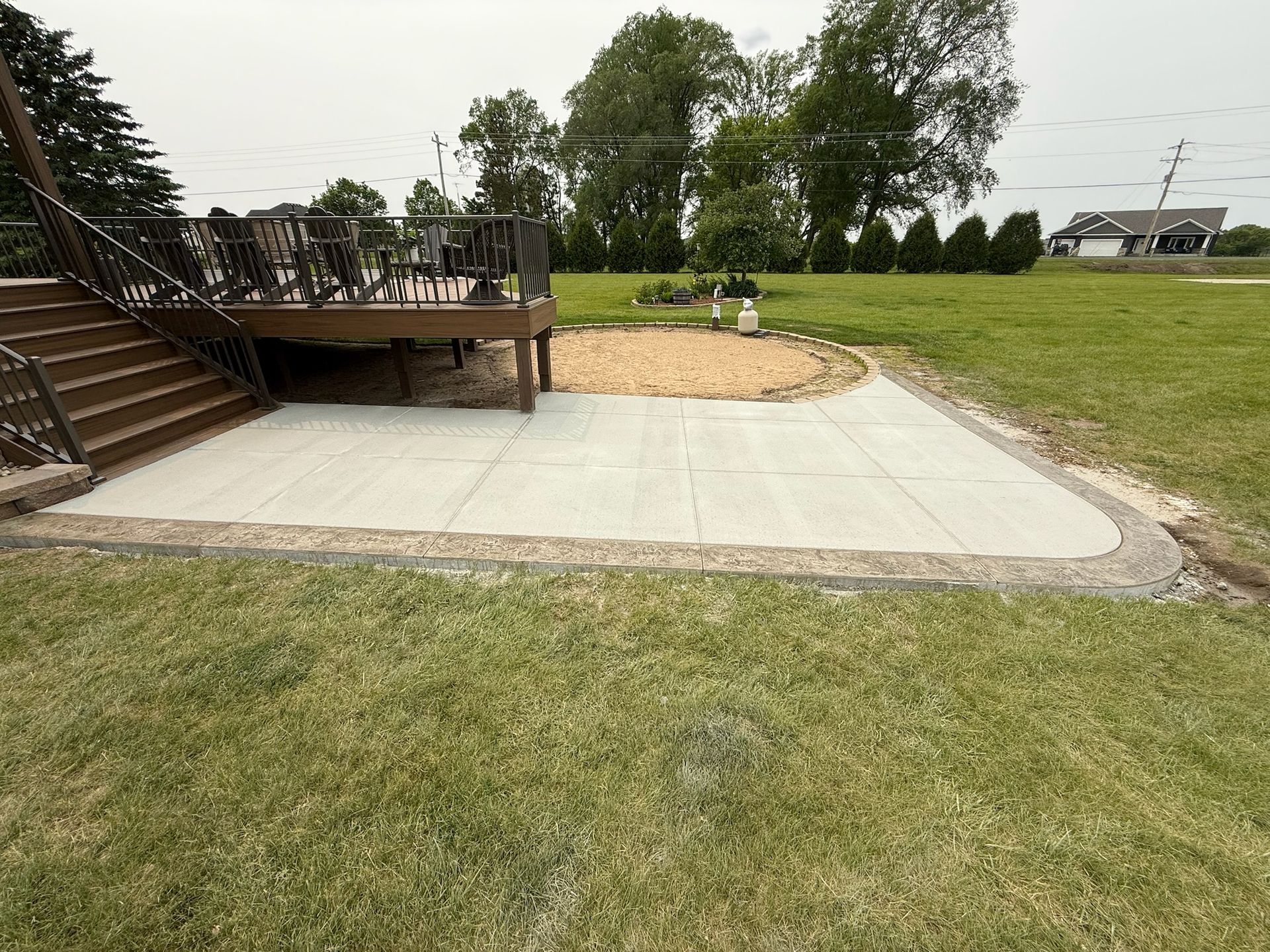 A concrete patio with a gravel border next to a wooden deck and staircase in a backyard with grass and trees.