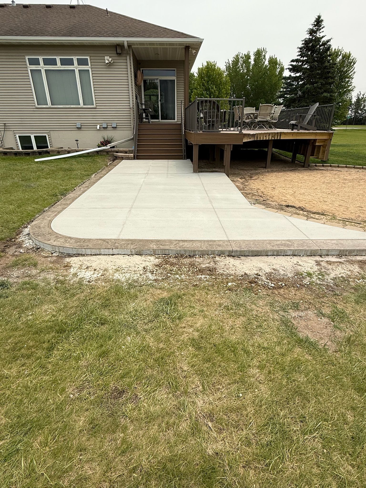 A house with a rear wooden deck and a newly poured concrete patio extension extending onto a grassy backyard.