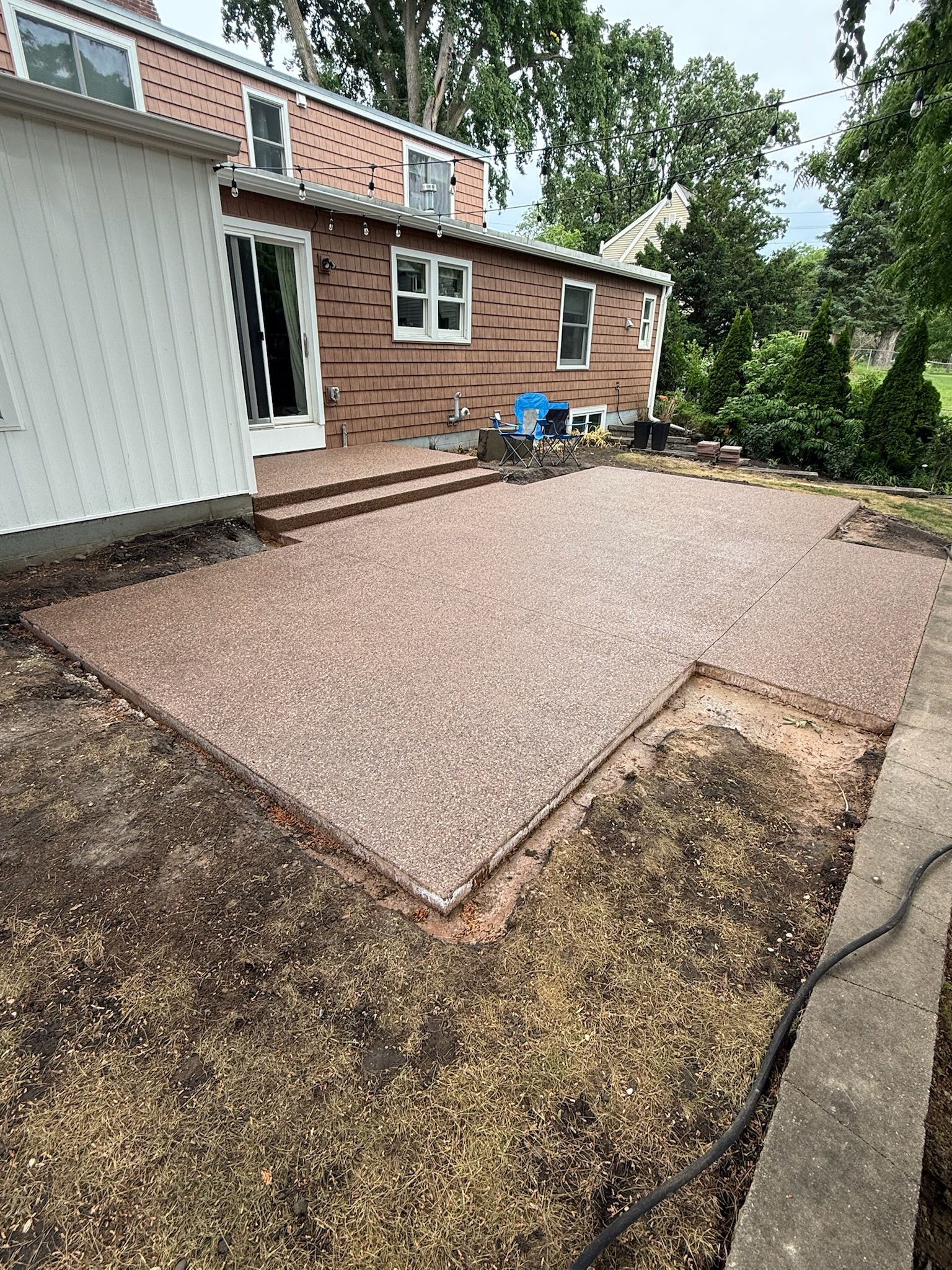 A newly installed textured brown stone patio in a backyard with a brick and white house, steps, and dirt landscaping.