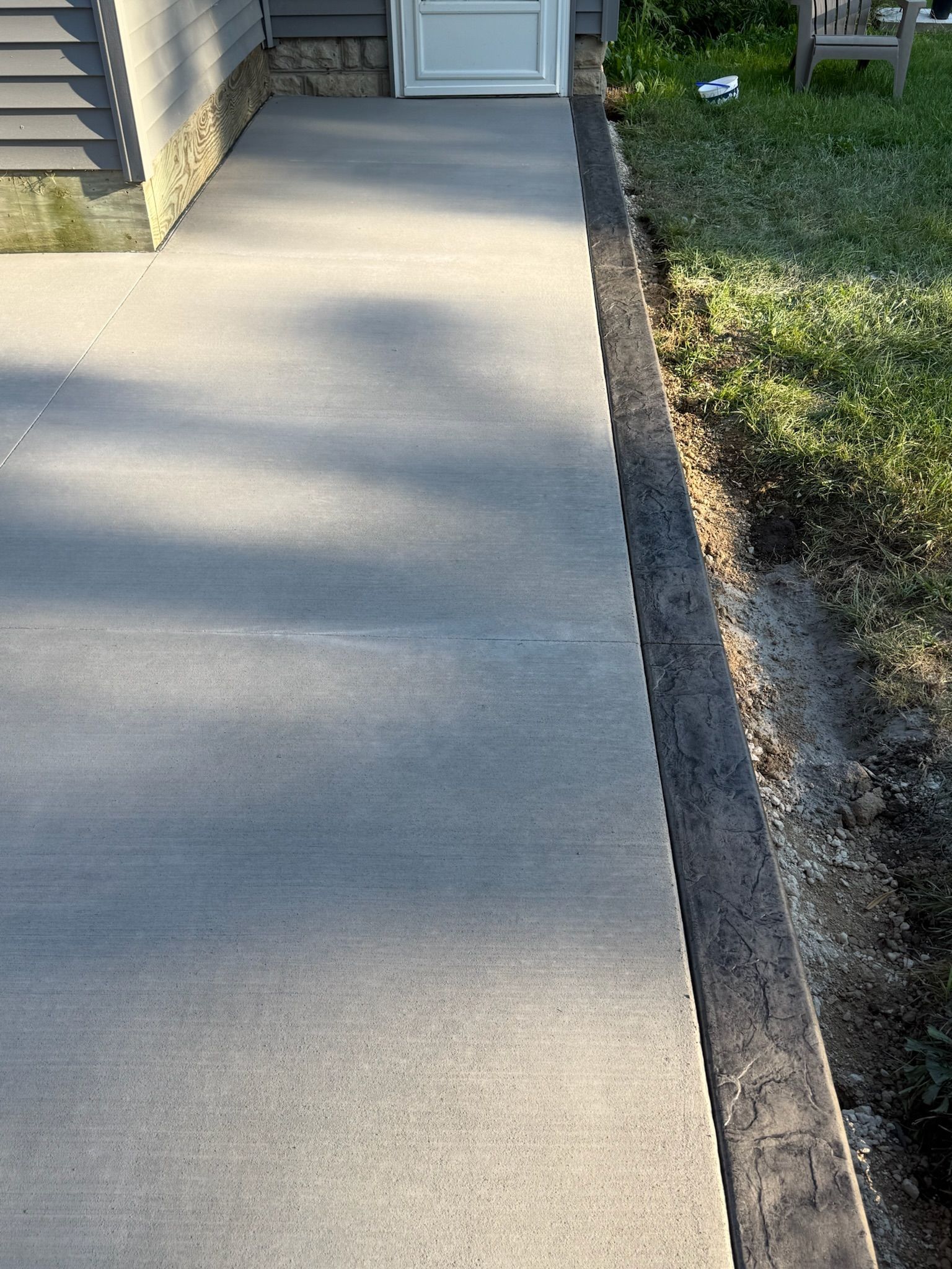 A concrete sidewalk path leading to a white door, with a dark wood edging running alongside the lawn.