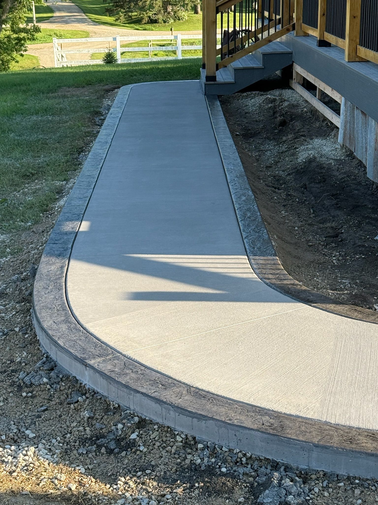 A newly poured concrete walkway with a dark stone border, curving alongside an elevated wooden deck in a grassy yard.