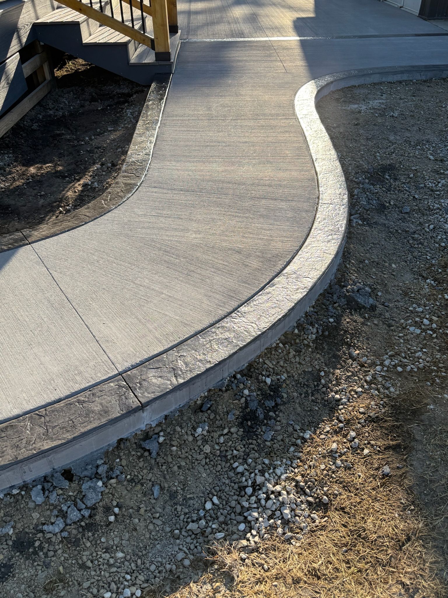 A curved concrete walkway lined with textured stone edging leads toward a wooden staircase outdoors.