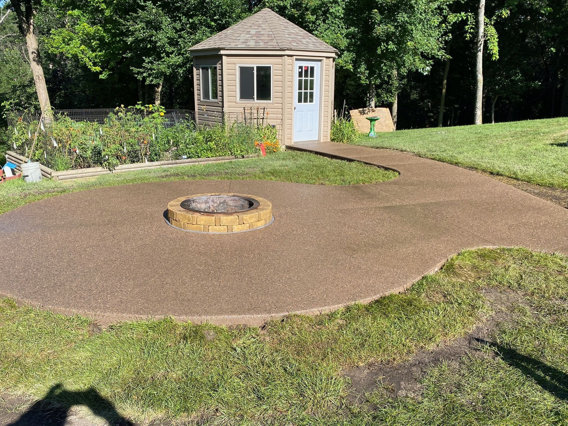 A backyard gravel patio featuring a circular stone fire pit, leading to a small tan shed with a blue door.