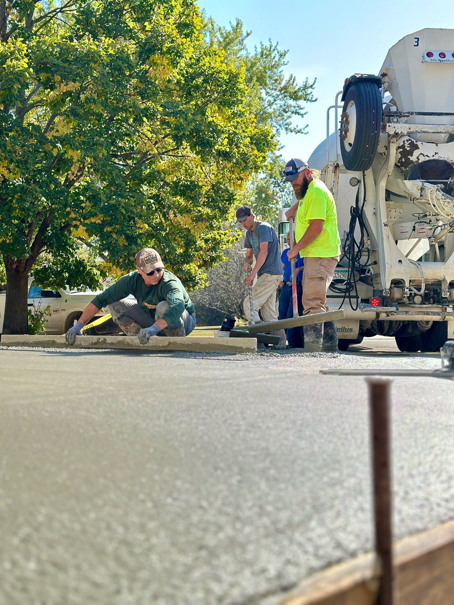 Three workers finish a fresh concrete pour next to a cement mixer truck on a sunny day.