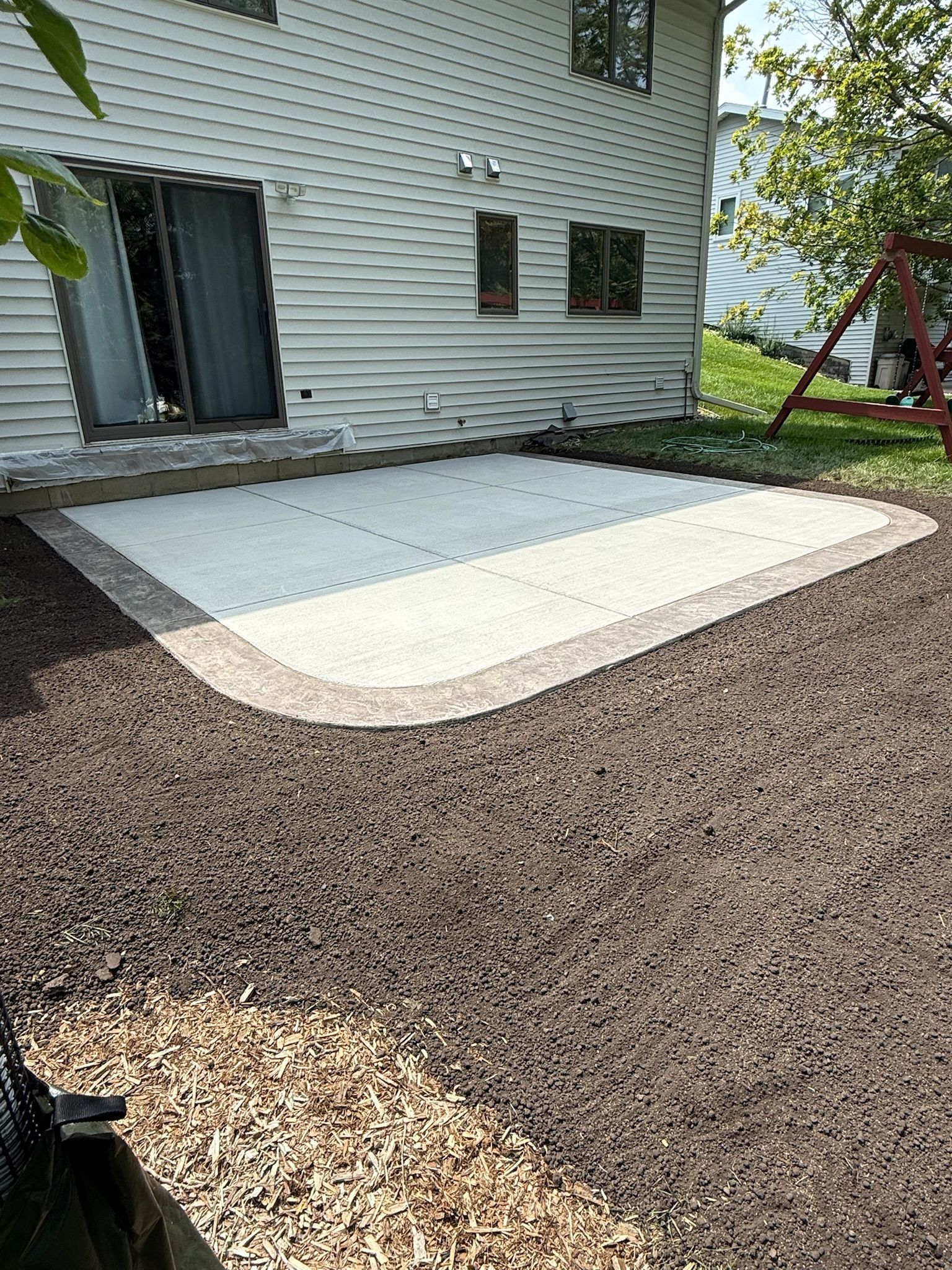 A new rectangular patio with light-colored center pavers and a dark border, set against the side of a beige vinyl house.