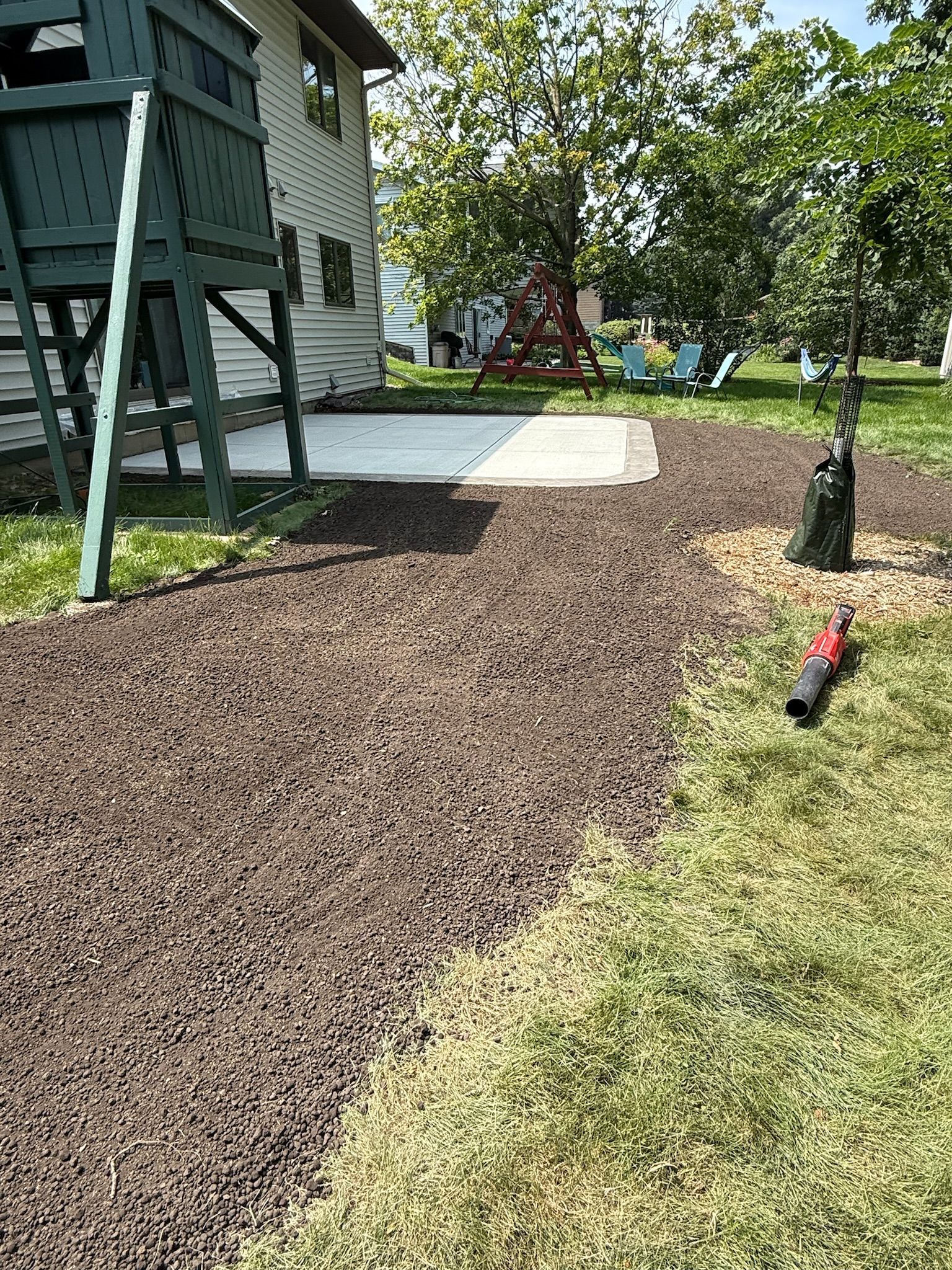 A backyard featuring a concrete patio, a green playground structure, and an area newly covered in dark brown mulch.