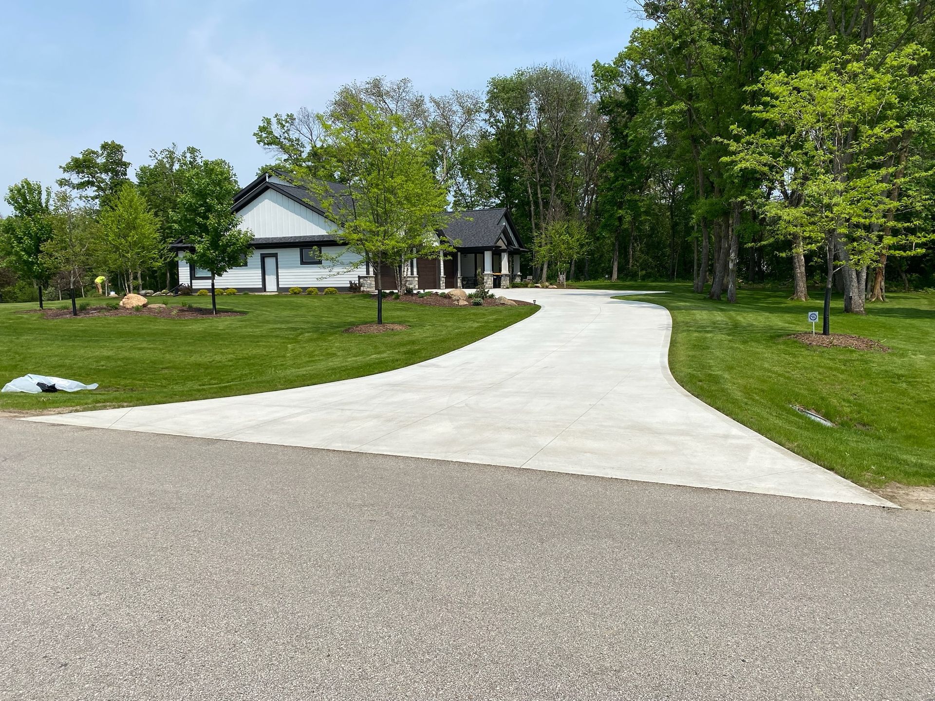 A new concrete driveway leads from an asphalt road to a modern white and dark-trimmed house surrounded by green lawns.