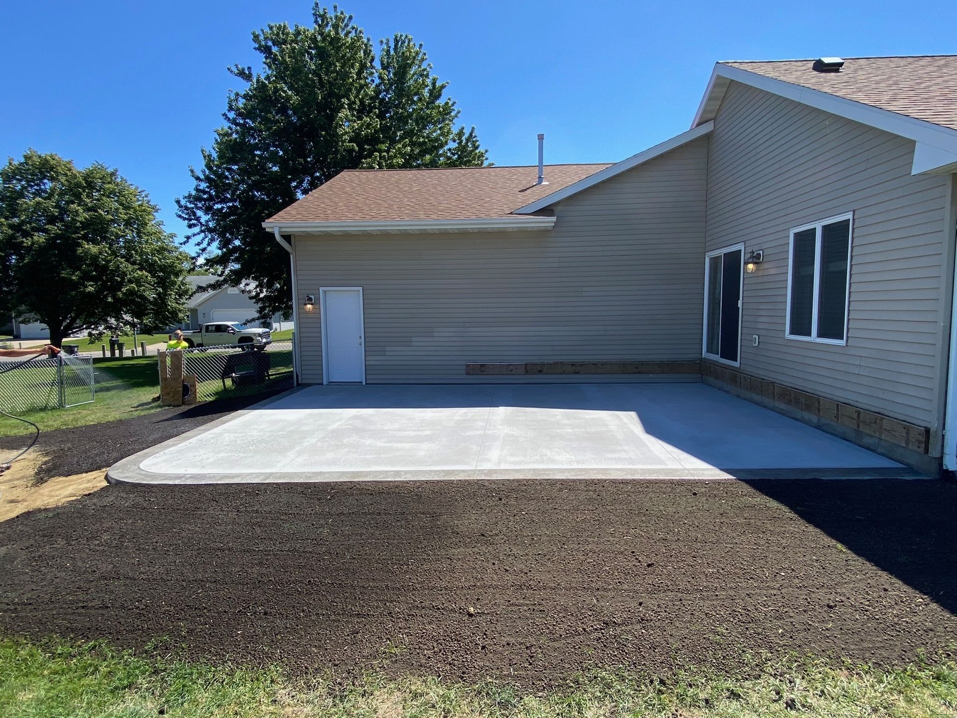 A newly poured concrete patio attached to the side of a tan-sided house, surrounded by fresh, dark soil under a blue sky.