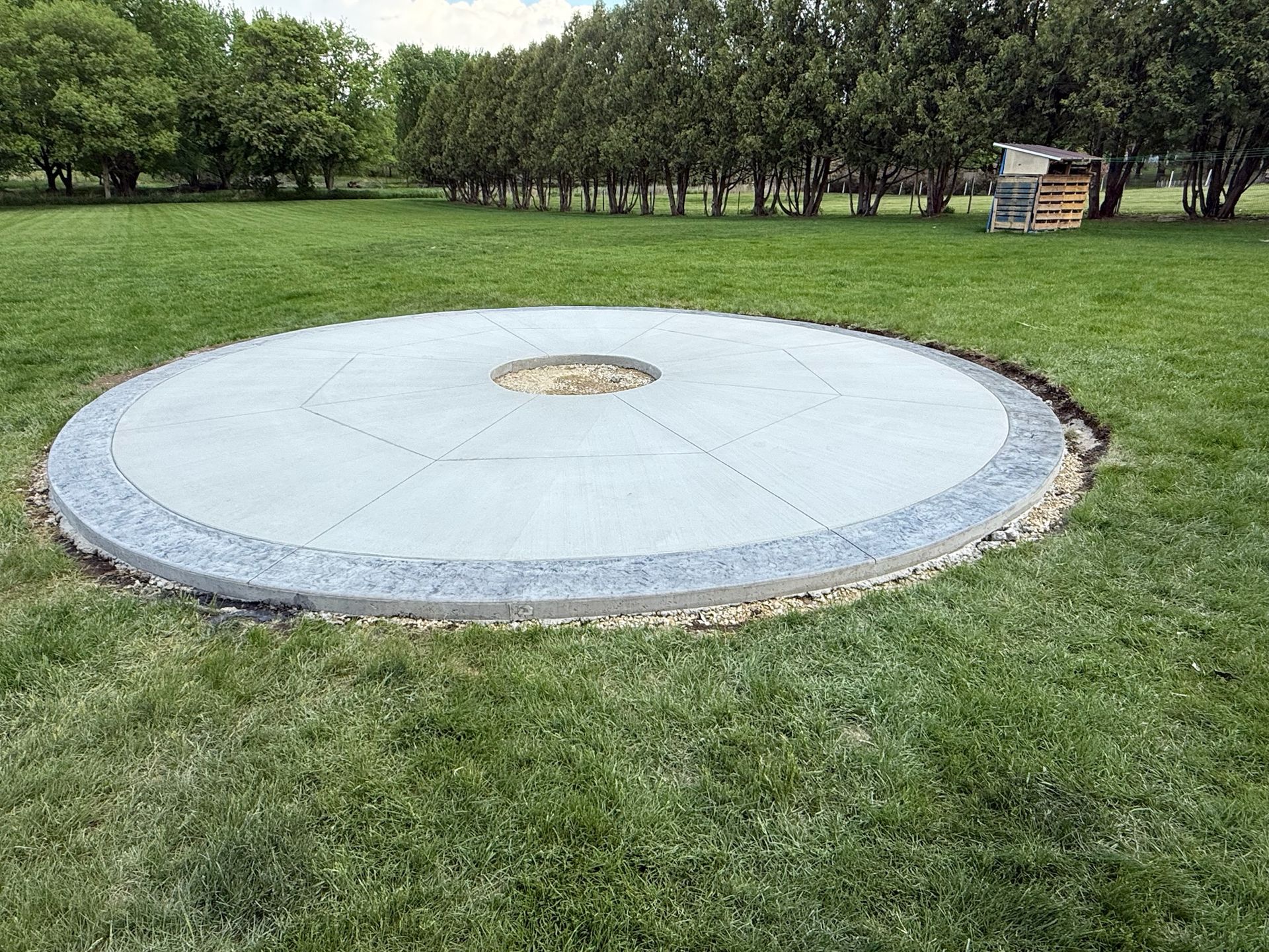 A round concrete patio with a central fire pit in a grassy field, bordered by a textured stone edge.