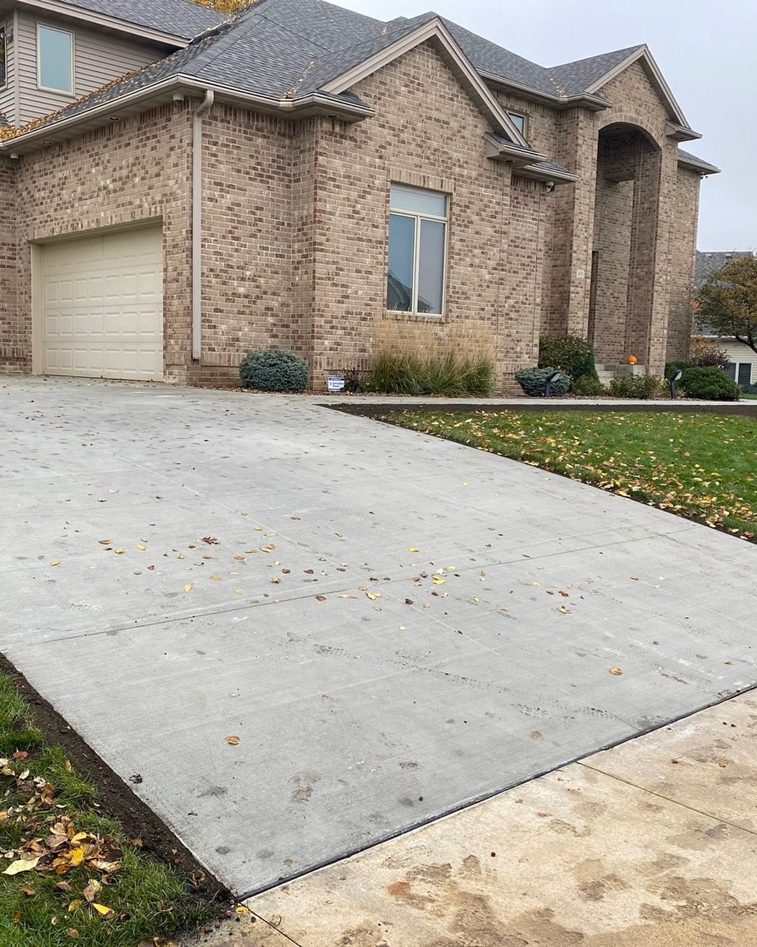 A beige brick house with a two-car garage and a concrete driveway, viewed from the street on an overcast day.
