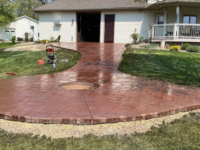 A red-toned, stamped concrete patio and walkway lead toward a house with a deck and garage.
