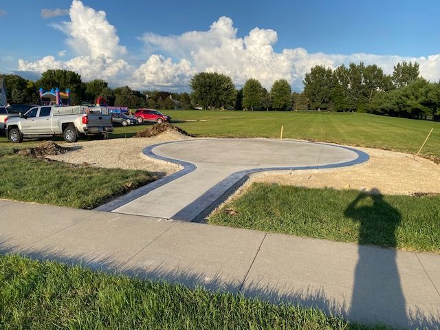 A newly poured concrete circular pad with a walkway connects to a sidewalk in a grassy field under a sunny sky.