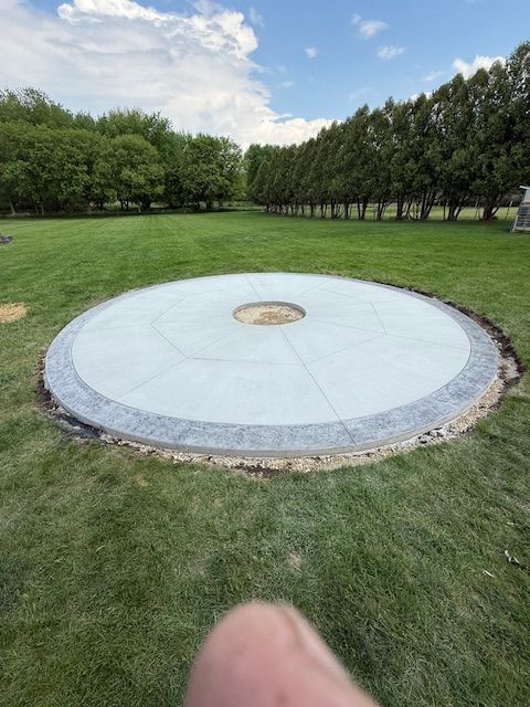 A circular concrete patio with a textured stone border and a small center fire pit opening, set in a grassy backyard.