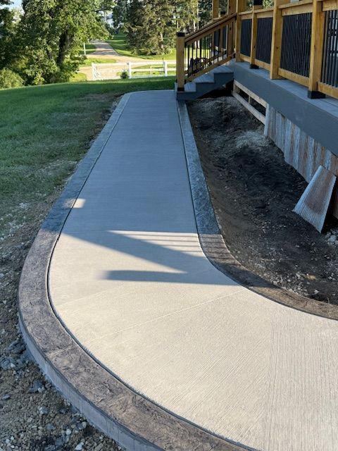 A newly poured concrete walkway with a dark, decorative border curves along the side of a wooden deck.