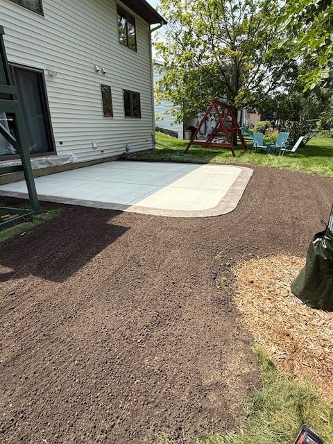 A newly installed concrete patio with a paved border set against the side of a light-sided house with a dirt yard.