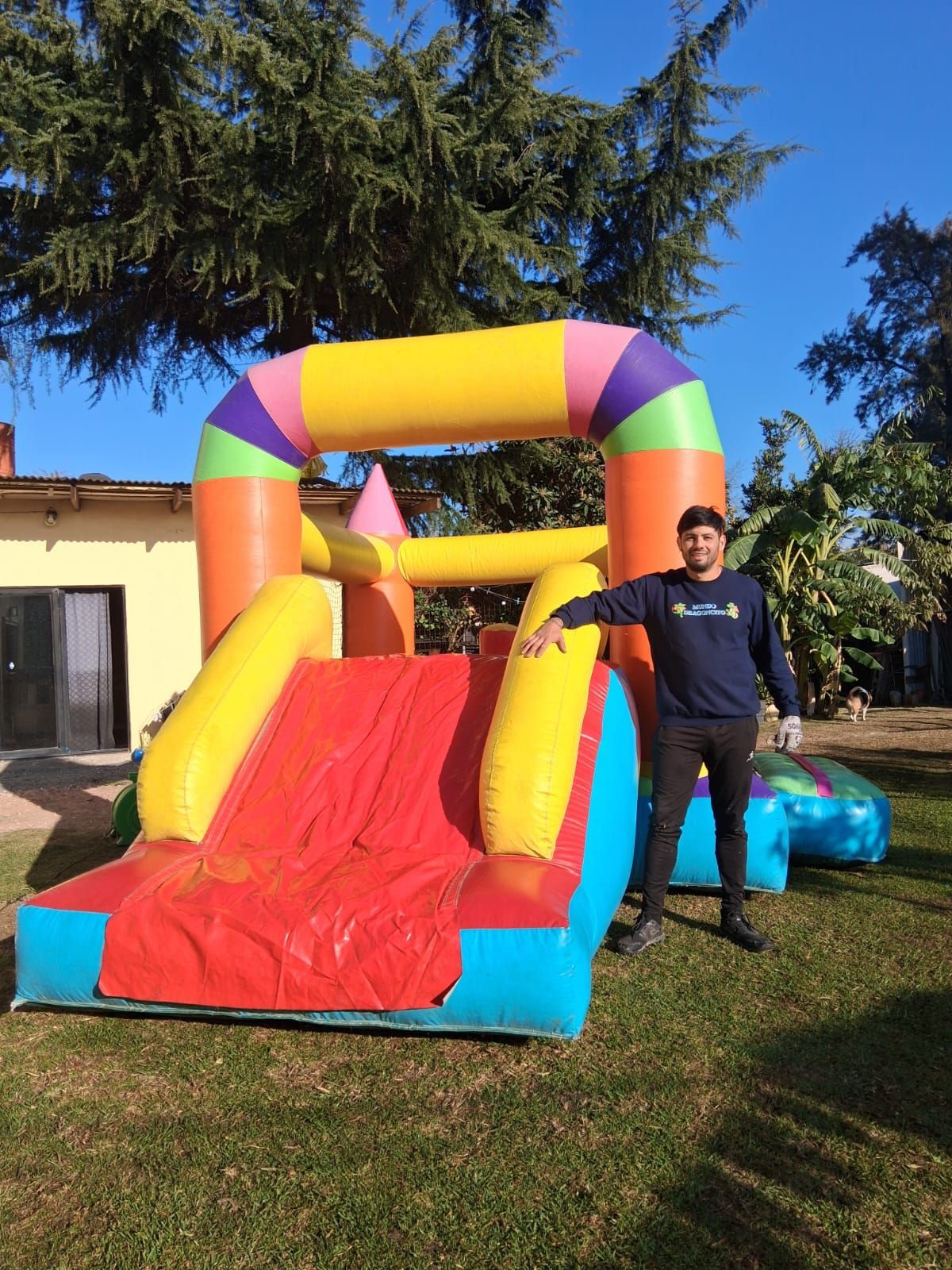 Tobogán inflable colorido al aire libre, con una persona de pie junto a él en un jardín con césped.