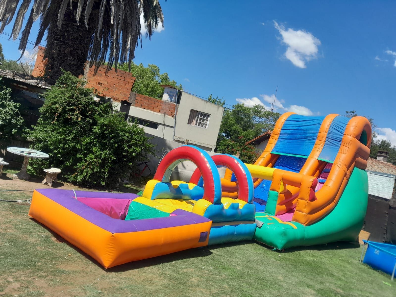 Tobogán acuático inflable colorido y castillo hinchable en un patio trasero soleado con palmeras.