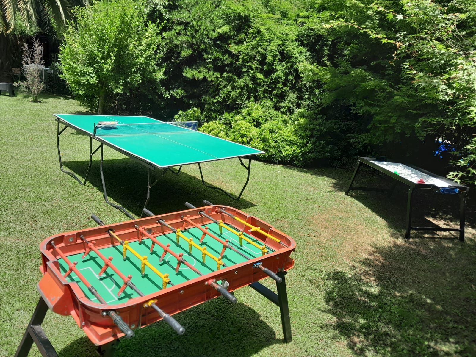 Dos mesas de futbolín al aire libre, sobre un césped, una naranja y otra verde, bañadas por la luz del sol.
