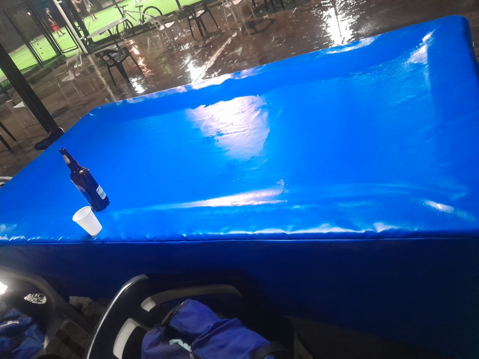 Mesa de billar azul bajo luces brillantes con una botella de taco en la esquina y lluvia afuera, cerca de las ventanas.