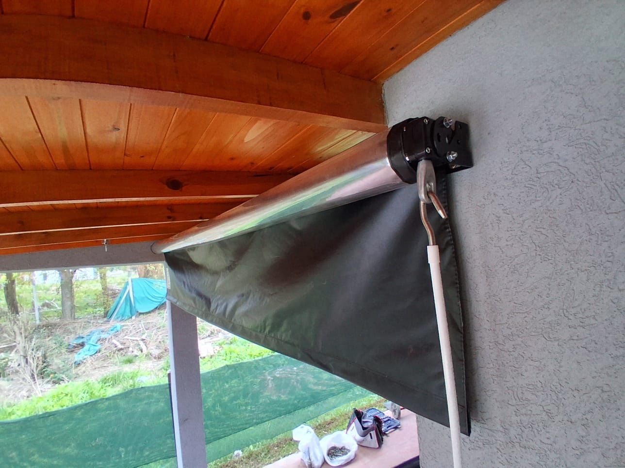 Toldo retráctil negro bajo un techo de madera, extendido sobre un porche con un jardín al fondo.