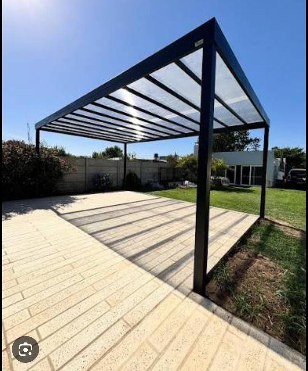 Patio trasero moderno con pérgola de metal negro y suelo pavimentado bajo un cielo azul.
