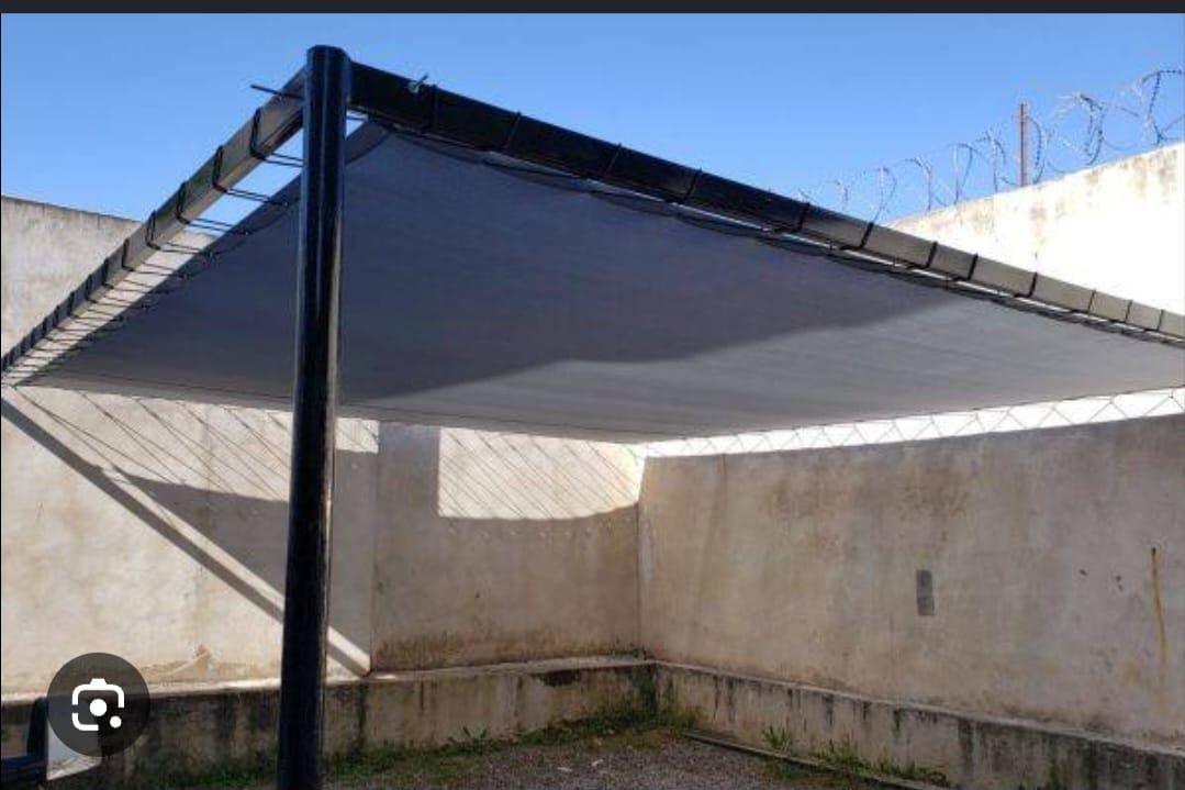 Un toldo de vela negra se extiende sobre un patio entre muros de hormigón y postes de metal.