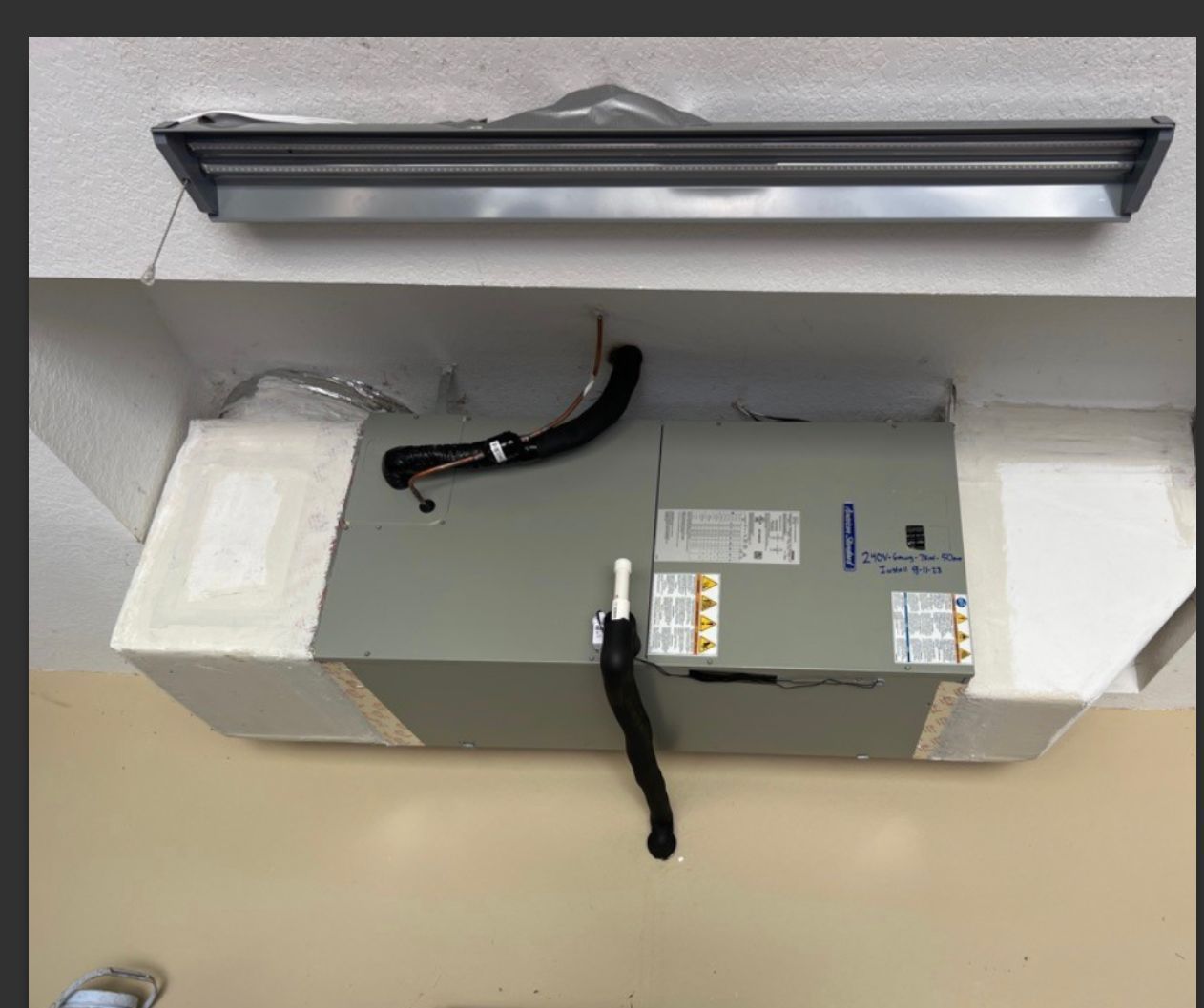 HVAC unit installed in a ceiling cavity, with ductwork and insulation. A fluorescent light fixture is above.