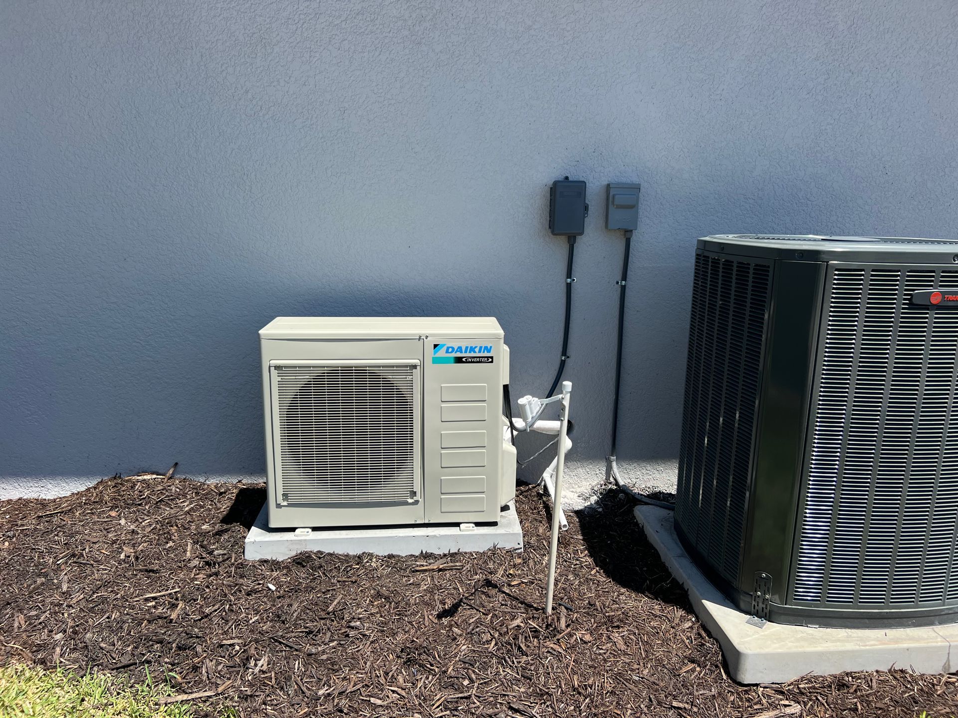 Two air conditioning units on concrete pads against a gray wall, on a bed of mulch.